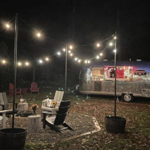 Airstream mobile bar parked next to outdoor fire pit with chairs at night